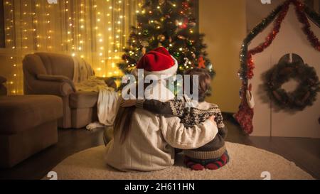 Rückansicht einer umarmenden Mutter mit Sohn in einem warmen Wollpullover, der neben dem glühenden Weihnachtsbaum sitzt Stockfoto