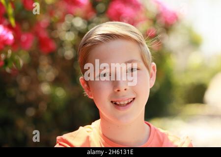 Porträt eines lächelnden, fröhlichen, blonden Jungen in orangefarbenem T-Shirt, mit Zahnspangen, im Freien unter sonnigen Bedingungen mit rosa Blüten im Hintergrund Stockfoto