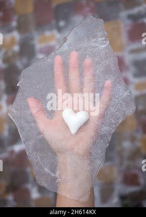 Ein weißes Keramikherz liegt auf der Hand, darüber ist ein Stück durchsichtiges Eis. Als Symbol der Liebe oder Trennung haben sich die Gefühle der Liebe abgekühlt. Liebe t Stockfoto