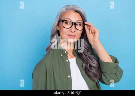 Foto von niedlichen Holder grau Frisur Dame suchen leeren Raum tragen Brillen Khaki Shirt isoliert auf blauem Hintergrund Stockfoto