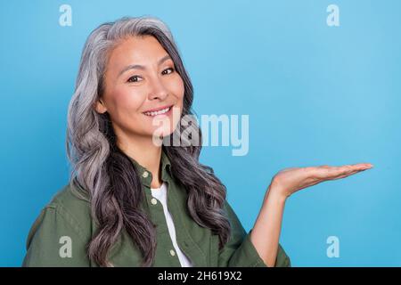 Foto von niedlichen Holder grau Frisur Dame halten leeren Raum tragen grünes Hemd isoliert auf blauem Hintergrund Stockfoto