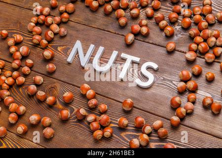 Das Wort Nüsse mit silbernen Buchstaben auf Holzbrett Hintergrund mit verstreuten Haselnüssen umgeben gelegt Stockfoto
