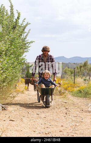 Ein junger Mann schiebt einen fröhlichen und aufgeregten Jungen, der in einer Schubkarre auf dem Gehweg auf der Farm sitzt Stockfoto