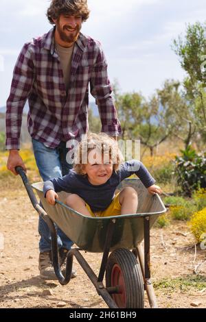 Glücklicher bärtiger Vater schiebt Sohn in Schubkarre auf Gehweg auf der Farm an sonnigen Tag sitzen Stockfoto