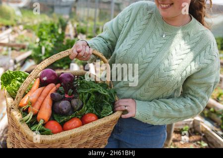 In der Mitte der lächelnden Bäuerin, die auf dem Bauernhof frisch geerntetes Gemüse im Korb hält Stockfoto