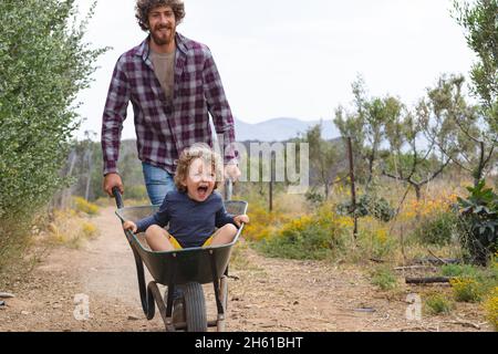 Junger Mann schiebt fröhlich und aufgeregt Sohn sitzen in Schubkarre auf Gehweg auf der Farm Stockfoto