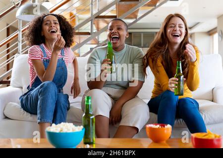 Multirassische Freundinnen mit Bier und Popcorn lachen, während sie zu Hause Fernsehen Stockfoto