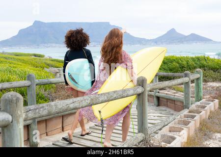 Freundinnen, die am Wochenende Surfbretter tragen, während sie am Strand auf der Promenade spazieren gehen Stockfoto