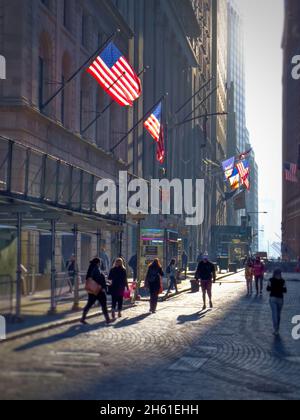 Silouette von Menschen in einer Kopfsteinpflasterstraße im unteren Manahattan in der Nähe der New Yorker Börse. Hinterleuchtete amerikanische Flaggen auf den Gebäuden Stockfoto