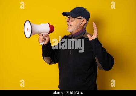 Reifer Mann trägt Kappe hält Megaphon isoliert über gelbem Hintergrund. Stockfoto
