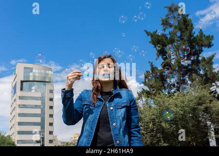 Porträt einer schönen hispanischen Frau, die an einem sonnigen Morgen Seifenblasen mitten in einem Park bläst Stockfoto