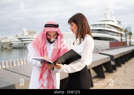 Ein lächelnder arabischer Geschäftsmann, der einen Vertrag mit einem Schreibergesang hält, während er mit einer jungen europäischen Geschäftsfrau im Hafen steht, ein arabischer Mann, der bei einem Treffen mit einem Geschäftspartner im Freien die Unterschrift auf die Dokumente legt Stockfoto