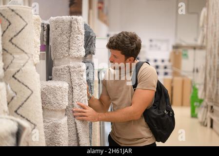 Fokussierter junger Mann mit Rucksack, der auf weichen grauen Teppich in Rolle schaut, während er in der Abteilung für Heimdekor einkauft Stockfoto