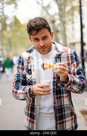 Lächelnder Mann, der süße Churros isst, während er mit einer Tasse heißer Schokolade in der Stadt steht und die Kamera anschaut Stockfoto