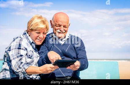 Glückliches Paar mit Senioren, das am Strand mit einem Tablet Spaß hat - Porträt von Mann und Frau, die mit moderner Technologie interagieren Stockfoto