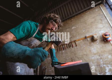 Bärtiger kaukasischer Schmied in Schutzkleidung Schmieden mit Hammer auf Amboss in der Industrie Stockfoto