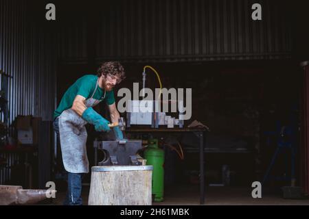 Kaukasischer Schmied in der Arbeitsschutzschmiede mit Hammer auf Amboss in der Industrie Stockfoto