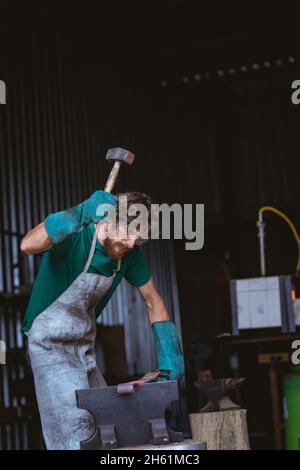 Kaukasischer bärtiger Schmied in Schutzkleidung Schmieden mit Hammer auf Amboss in der Industrie Stockfoto