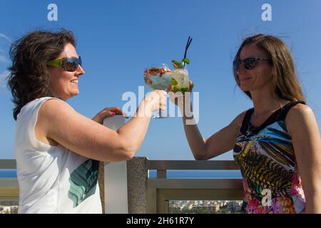 Ein paar Mädchen stoßen auf der Dachterrasse mit Cocktailgetränken an. Lächelnde Frauen mit Sonnenbrillen auf Party-Feier. Junge Damen, die Spaß haben Stockfoto