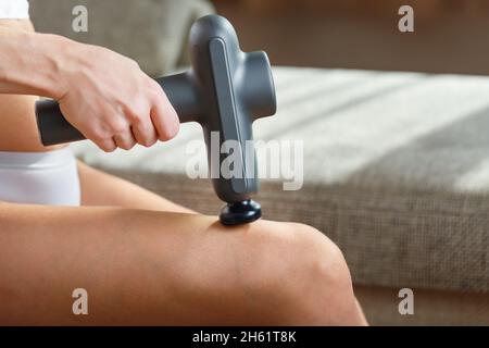 Selbstmassage der Frauenbeine mit einer perfektionierenden Pistole zu Hause. Schockmassage zur Wiederherstellung der Faszien- und Triggerpunkte Stockfoto