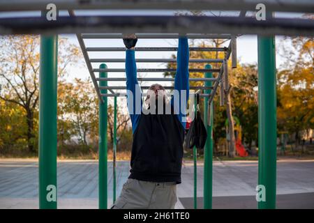 Der stark grauhaarige Mann mit Dreadlocks ordnet seine Hände an der horizontalen Affenbar im Freien neu. Klettergerüst für sportliche Aktivitäten im Freien. Erwachsener Stockfoto