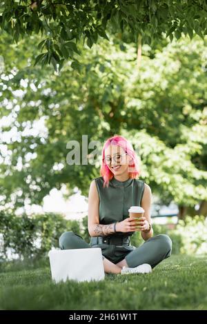 Glückliche Frau mit rosa Haaren und Brillen halten Papierbecher und Blick auf Laptop auf Gras Stockfoto