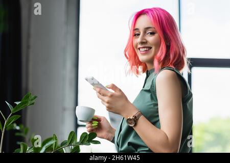 Fröhliche Geschäftsfrau mit rosa Haaren und Piercing hält Tasse Kaffee und Smartphone Stockfoto