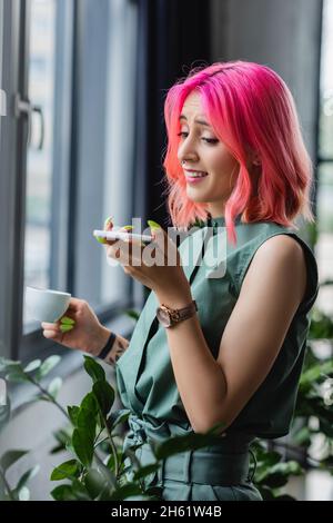 Fröhliche Geschäftsfrau mit rosafarbenem Haar und Piercing, die eine Tasse Kaffee hält, während sie die Sprachnachricht auf dem Smartphone aufzeichnet Stockfoto
