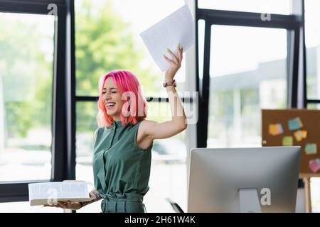 Fröhliche Geschäftsfrau mit rosafarbenem Haar und durchdringender Aufbewahrung Ordner mit Dokumenten im Büro Stockfoto