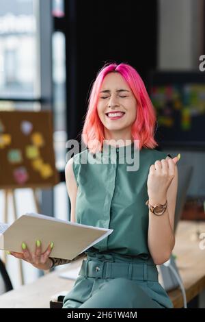 Fröhliche Geschäftsfrau mit rosa Haaren und durchdringenden Ordner im Büro halten Stockfoto