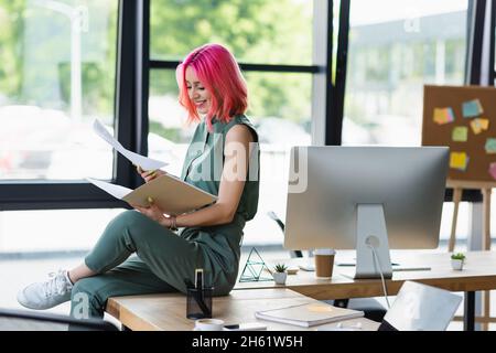Fröhliche Geschäftsfrau mit rosafarbenem Haar und durchdringenden Händen Ordner und Dokumente im Büro Stockfoto