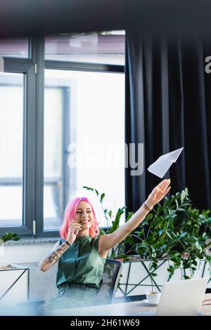 Positiver Manager mit rosa Haaren, der auf dem Smartphone spricht, während er ein Papierflugzeug wirft Stockfoto