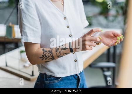 Beschnittene Ansicht einer tätowierten Geschäftsfrau, die im Büro Desinfektionsmittel auf die Hand sprüht Stockfoto