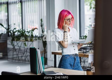 Nachdenkliche junge Geschäftsfrau mit rosafarbenem Haar, die ihren Laptop im Büro hält Stockfoto