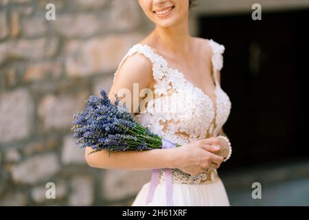 Sibenik, Kroatien - 05.06.17: Lächelnde Braut in einem weißen Spitzenkleid hält einen Lavendelstrauß auf ihrem Ellbogen und steht an einer Steinmauer Stockfoto