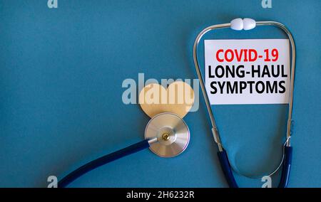 COVID-19 Symbol für langstreckige kovide Symptome. Weiße Karte mit den Worten Covid-19 Langstrecken-Symptome. Holzherz, Stethoskop, blauer Hintergrund, Kopierraum. M Stockfoto