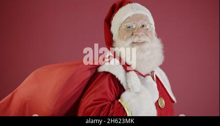 Echter Weihnachtsmann mit rotem Hintergrund, mit Brille, Handschuhen und einem Hut auf der Seite. Stockfoto