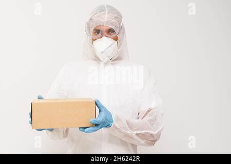 Ein Mann trägt Schutzkleidung mit Gesichtsmaske, Handschuhen und Schutzanzug, der eine Schachtel hält Stockfoto