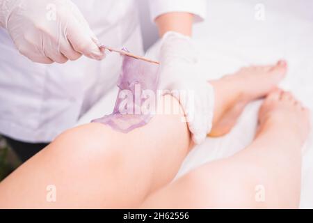 Kosmetologe in der Beauty-Klinik führt ein Enthaarungsverfahren für die Entfernung von wachsenden Haaren aus den Beinen mit Wachs violett Zuckerpaste, Schönheit und Körper Stockfoto