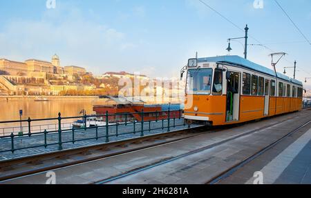 Die gelbe Retro-Straßenbahn am Belgrader Kai gegen die Donau und die Budaer Burg, im Hintergrund bei hellem Sonnenlicht gesehen, Budapest, Ungarn Stockfoto