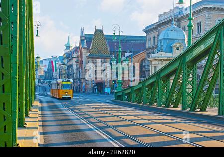 Die landschaftlich reizvolle gelbe Straßenbahn fährt entlang des Fovam-Platzes zur Freiheitsbrücke, Budapest, Ungarn Stockfoto