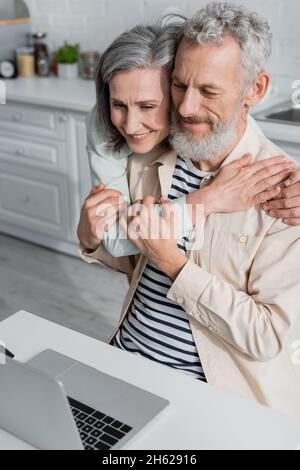 Reifes Paar umarmt sich in der Nähe von Laptop in der Küche zu Hause Stockfoto