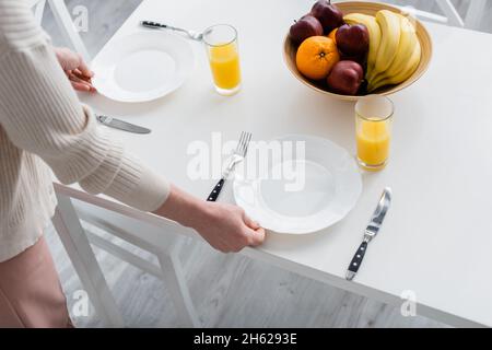 Beschnittene Ansicht einer Frau, die Teller auf den Tisch neben Orangensaft und Obst in der Küche legte Stockfoto
