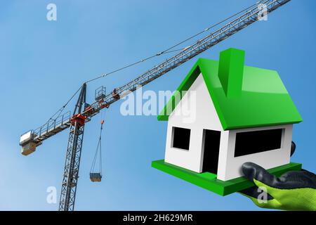 Nahaufnahme einer Hand mit schützenden Arbeitshandschuhen, die ein kleines Modellhaus mit einem Kran gegen einen klaren blauen Himmel hält. Konzept der Bauindustrie. Stockfoto