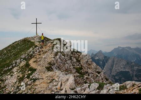 Junge Frau auf der Wanderung vom Gipfel der Brunnsteinspitze (2197 Meter) ins Tal, von vorne, Gipfelkreuz im Hintergrund, österreich, tirol, scharnitz, Naturpark karwendel, Stockfoto