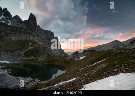 Eine einzelne, kaum sichtbare Person steht am Rande eines Bergsees und blickt auf den Sonnenuntergang in der rauen Landschaft, montenegro, albanien, prokletije Nationalpark Stockfoto