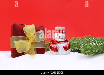 Schneemann mit Geschenk-Box in Schnee und grünen Fichtenzweigen auf rotem Hintergrund. Weihnachten, Neujahr. Speicherplatz kopieren Stockfoto