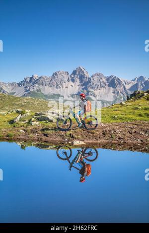 Mann, 22 Jahre alt, Radfahrer mit E-Bike in der Nähe eines alpinen Sees, Radfahrer im Wasser eines Bergsees reflektiert, Berge im Hintergrund. Laresei, falcade, belluno, veneto, italien Stockfoto