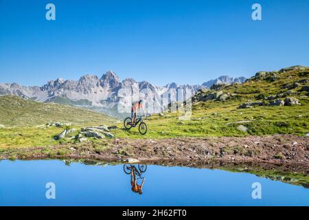 Mann, 22 Jahre alt, Radfahrer mit E-Bike in der Nähe eines alpinen Sees, Radfahrer im Wasser eines Bergsees reflektiert, Berge im Hintergrund. Laresei, falcade, belluno, veneto, italien Stockfoto