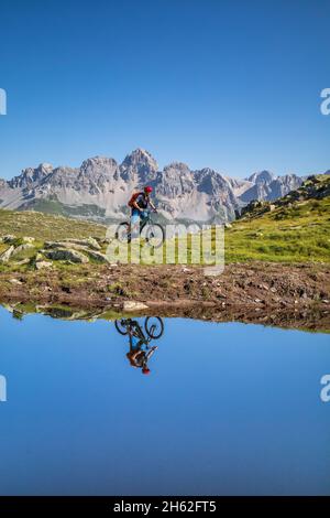 Mann, 22 Jahre alt, Radfahrer mit E-Bike in der Nähe eines alpinen Sees, Radfahrer im Wasser eines Bergsees reflektiert, Berge im Hintergrund. Laresei, falcade, belluno, veneto, italien Stockfoto